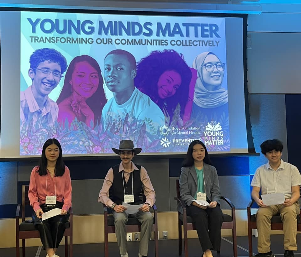 The image shows four young adults seated in front of a large projection screen displaying the words "YOUNG MINDS MATTER - TRANSFORMING OUR COMMUNITIES COLLECTIVELY." The backdrop features colorful portraits of diverse young individuals, aligning with the event's theme. The first person on the left is a woman wearing a pink blouse, the second person is a man with a mustache and a hat, the third is a woman in a gray blazer, and the fourth is a man in a light-colored shirt. They all hold papers and appear to be part of a panel or presentation, possibly at a conference or seminar focused on youth and mental health, as indicated by the logos of the Hogg Foundation for Mental Health and Prevention Institute on the screen. The setting suggests an academic or professional environment.