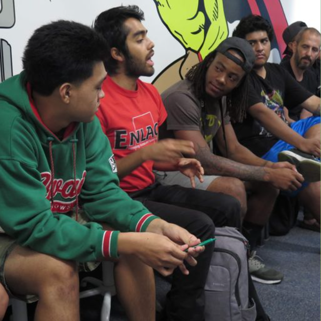 The image depicts a group of young men sitting and engaging in a discussion. The man in the foreground is wearing a green hoodie with the word "Coca-Cola" in white lettering. Beside him, a man in a red shirt with the text "ENLACE" is animatedly speaking and gesturing with his hand, indicating an active conversation. A man with dreadlocks, wearing a grey sleeveless shirt, is listening intently, and another in a blue shirt appears to be observing the discussion. In the background, there's a man with a beard, partially obscured. They are seated against a backdrop featuring stylized graffiti art, adding a vibrant and creative atmosphere to the setting. The image captures a moment of community engagement or a workshop setting, with a focus on dialogue and exchange of ideas.