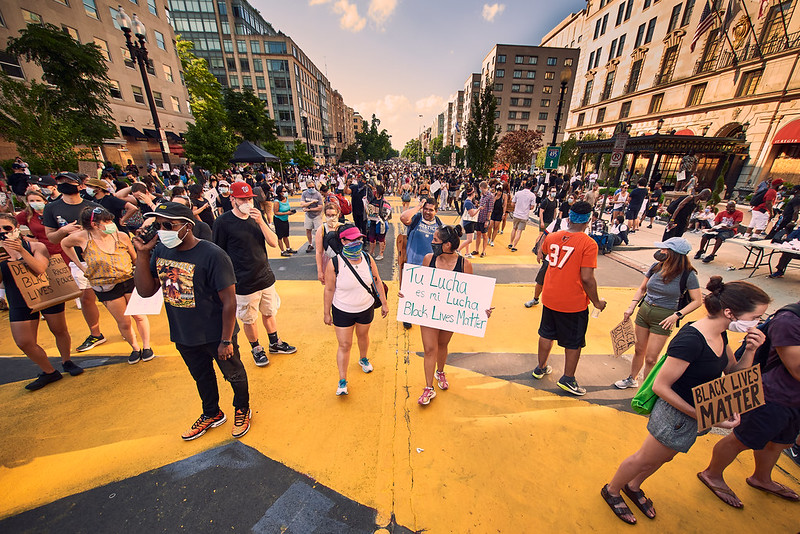 The image is a vibrant and powerful snapshot of a peaceful protest. A diverse crowd of demonstrators, many wearing face masks, gather on a street that has been painted with the words "Black Lives Matter" in large, bold letters. A woman in the center holds a sign that reads "Tu Lucha es mi Lucha Black Lives Matter," signifying solidarity across cultures in the fight against racial injustice. Individuals in the crowd are holding up their fists and signs, conveying a sense of unity and purpose. The setting is urban, with buildings lining the street, and the protest appears to be well-attended, indicating a strong community response to social issues. The photograph captures a moment of activism and communal support for the Black Lives Matter movement.