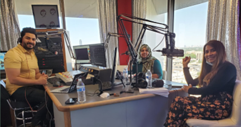 The image is of three people inside a radio studio, smiling and looking towards the camera. On the left is a man with a beard, wearing a yellow shirt, sitting in front of a microphone and a laptop, with headphones around his neck. In the center is a woman wearing a hijab and a teal top, seated at the control desk with microphones and monitors in front of her. On the right, there is another woman with long hair, wearing a cream top and floral skirt, sitting on a stool with a microphone in front of her. Behind them, there is a window showing a cityscape, and the room is well-lit, suggesting it is daytime. The overall mood is cheerful and professional.