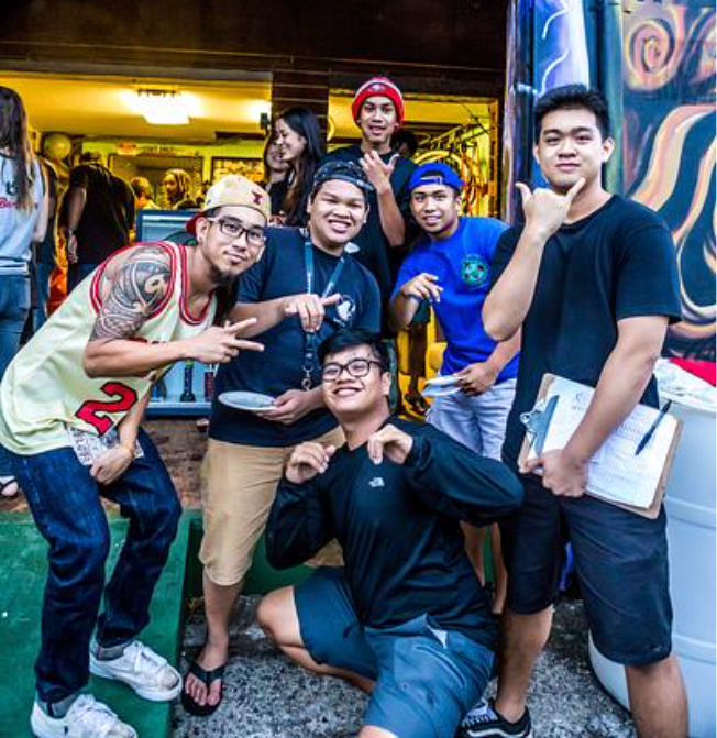 The image shows a joyful group of young adults posing for a photo. The scene appears to be an outdoor event or gathering. One individual is crouched in front with a big smile, making a playful gesture with his hands, while the others stand behind him, each striking a casual pose and displaying various expressions of happiness and camaraderie. They are all dressed in casual streetwear, and some wear hats or carry items like a clipboard, suggesting they might be part of an event staff or volunteers. The background includes a mural, adding an artistic and vibrant atmosphere to the scene. Overall, the image conveys a sense of fun, community engagement, and youthfulness.