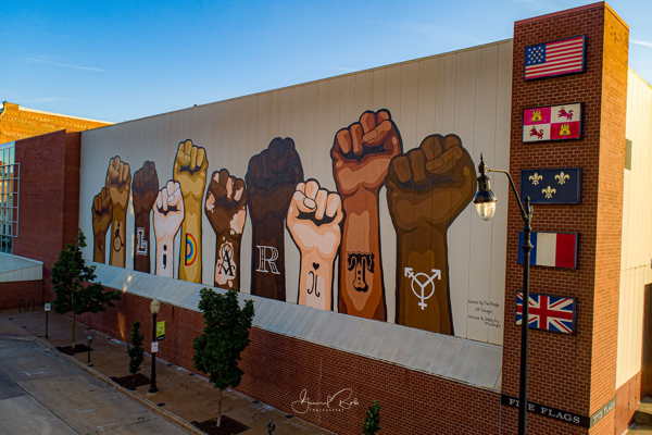 The image shows a mural of raised fists in different skin tones, each adorned with a letter to spell out "SOLIDARITY." Beside the fists are various flags, including the United States flag, indicating international unity or support for a cause. The mural is on a large building wall, viewed under a clear sky, and suggests themes of diversity, unity, and collective strength.