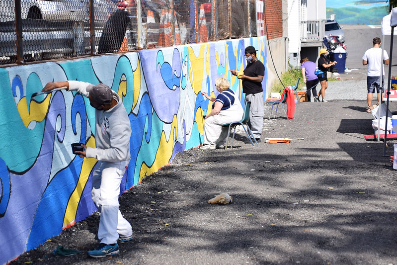 The image shows a group of people engaged in a community mural painting activity. They are outdoors, working on a wall that already features a partially completed abstract design with blue and yellow elements. Some individuals are standing while painting, and others are seated. The painters are wearing casual clothing, and some have protective clothing like overalls. The atmosphere appears to be casual, with some people focused on their painting and at least one person checking a phone. The setting includes a paved street and a chain-link fence in the background, hinting that this might be an urban environment. The weather seems to be clear, and shadows indicate it might be midday.