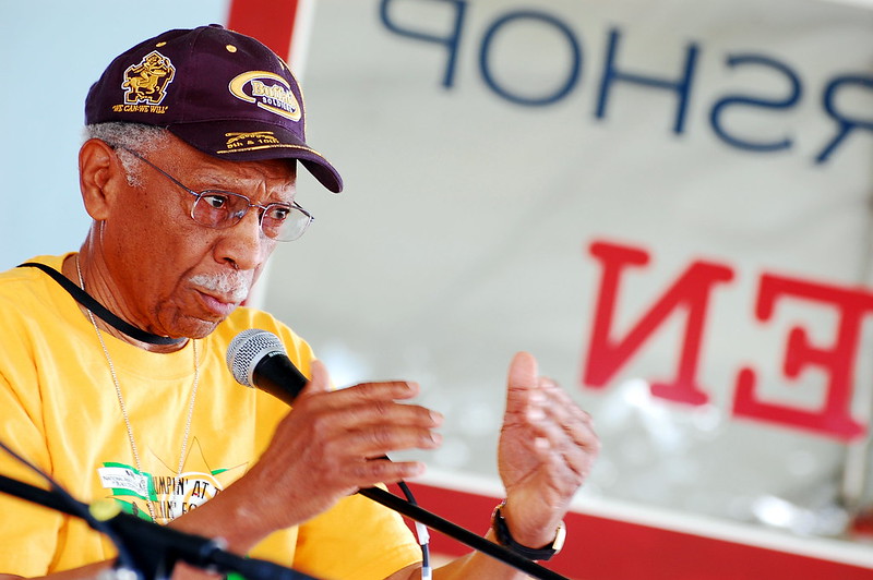 The image features an elderly Black man speaking into a microphone. He is wearing glasses, a baseball cap with an insignia, and a yellow T-shirt with some text and graphics visible. There is a banner in the background with reversed lettering, suggesting the word "SPEECH," indicating that the man is likely at a public speaking event or rally. His expression is focused and seems to convey a sense of seriousness or passion about the topic he is discussing.