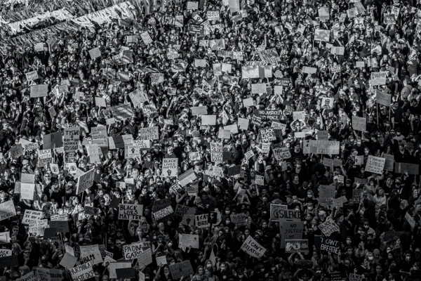 The image is a black and white photograph capturing a dense crowd of protesters. The mass of people is holding up various signs, creating a sea of placards that suggest a large-scale demonstration or public event focused on a cause or multiple issues. The absence of color emphasizes the unity and collective voice of the participants. The photo conveys the magnitude and intensity of public demonstrations and the power of collective action.