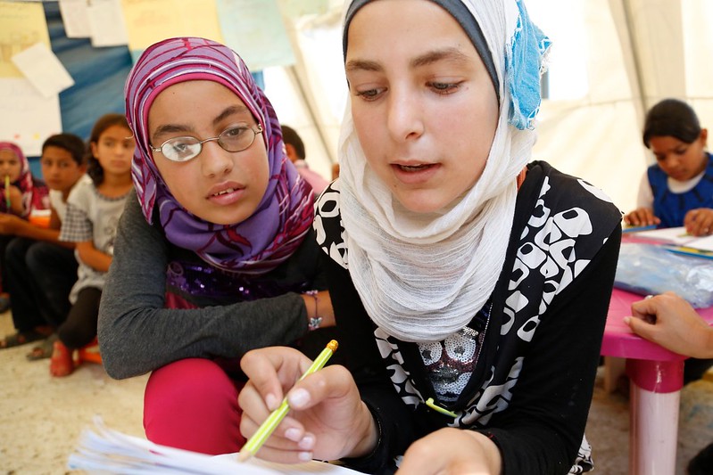 The image shows two young girls engaged in an educational activity. The girl in the foreground is wearing a vibrant purple and pink hijab and glasses, and is attentively watching as the girl in the background, wearing a white and blue hijab, writes in a notebook. They both appear to be in a classroom setting, indicated by other children and educational materials in the background. The focus on the two girls suggests a learning environment that fosters interaction and guidance, with a sense of concentration and dedication to the task at hand.