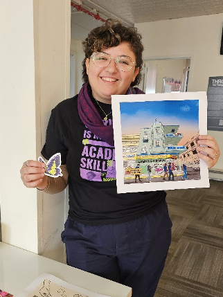 Smiling person holds colorful artwork depicting a building and a butterfly sticker while standing indoors.