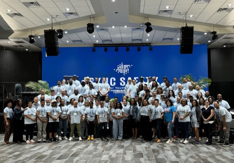 Large group of attendees posing on a stage before a blue SYNC SAFETY backdrop, smiling toward the camera.