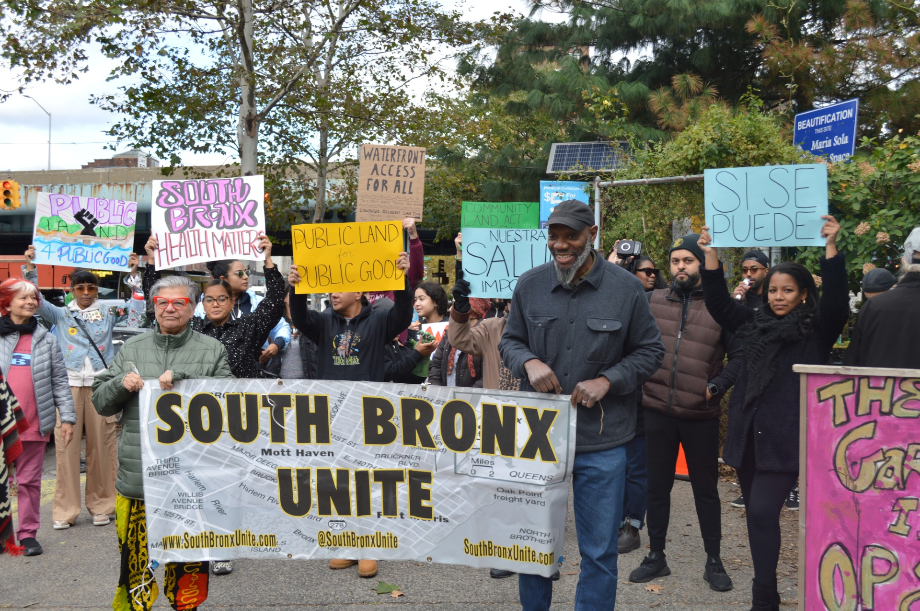 A diverse group of people at a community rally holding signs advocating for public land, health, and environmental justice. The signs read messages like "South Bronx Health Matters," "Public Land for Public Good," "Waterfront Access for All," and "Sí Se Puede." At the front, two individuals hold a large banner that says "South Bronx Unite." The setting includes trees, a beautification sign, and solar panels in the background.