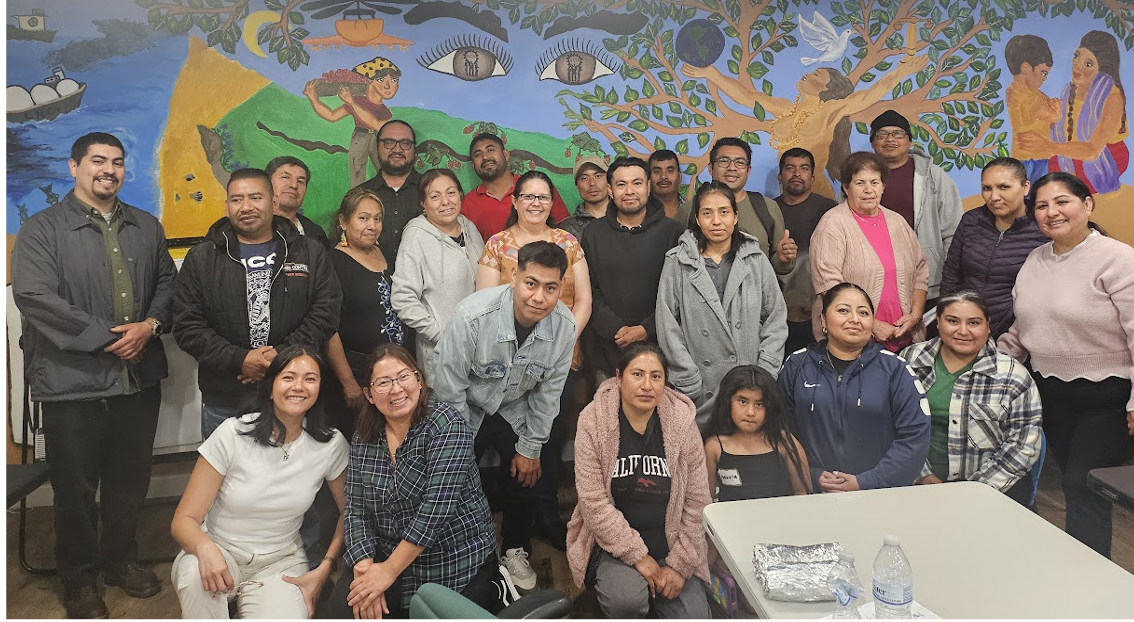 A diverse group of adults and one child pose together indoors for a group photo. They are standing and seated in front of a large, colorful mural that includes trees, people, eyes, and symbolic imagery related to community and nature. The group appears relaxed and smiling, suggesting a community gathering or workshop. A table with a water bottle and wrapped food sits in the foreground.