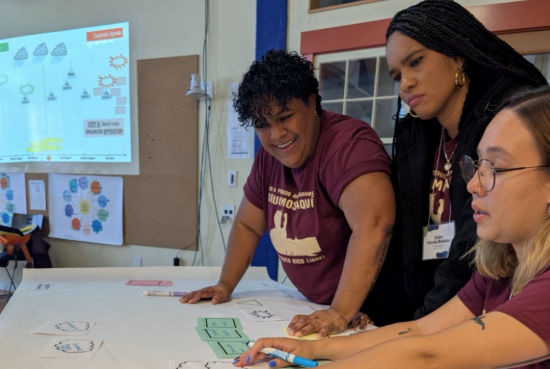 Three community members collaborate at a table reviewing materials, with a presentation slide visible in the background