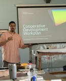 Man presents Cooperative Development Workplan slide to small group in meeting room while participants take notes at table.