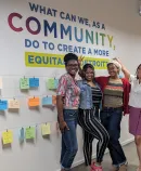 four cheerful women in front of a mural reading "What Can We, as a Community, Do to Create a More Equitable Detroit?"