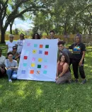 Group of community members outdoors holding a quilt with colorful squares