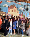 a group of cheerful, diverse individuals gathered for a photo in front of a whimsical mural depicting a town scene