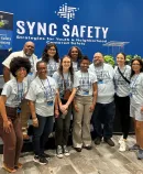 A group posing in front of a blue 'SYNC SAFETY' backdrop