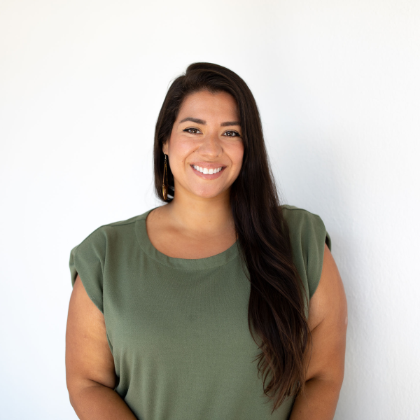 Smiling woman with long dark hair in a green top, posed against a bright white wall for a professional portrait.