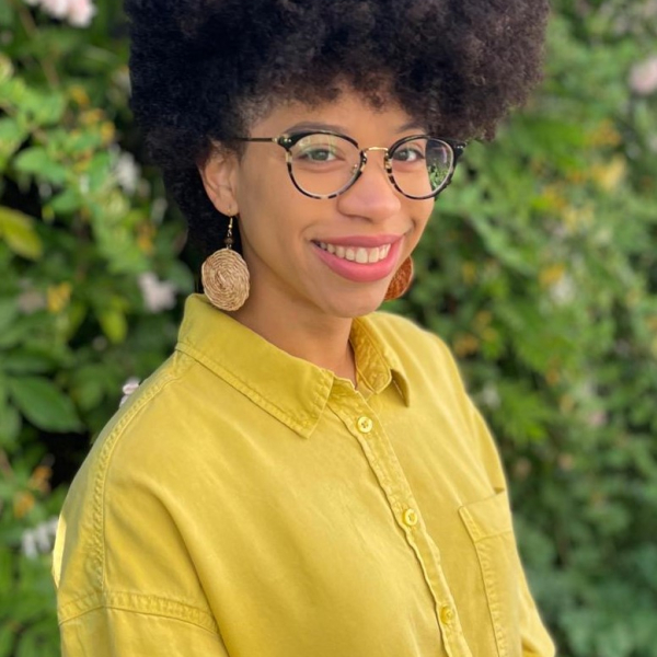 A person with medium-brown skin and a large, rounded curly afro stands outdoors in front of dense green bushes with pink flowers. They wear round glasses, large light-colored circular earrings, and a bright yellow long-sleeve button-down shirt. They are smiling gently and looking toward the camera with their body angled slightly to the side.