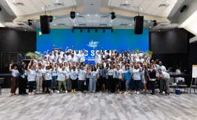 Large group of conference attendees in matching blue shirts pose with raised fists on a stage beneath a large blue screen.