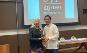 Two people smile holding a bronze statuette award beneath a screen reading "SJF 40 Years Rooted in Love" at the award event.