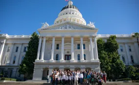 Large group of advocates, some in white coats, gathered on the steps of the California State Capitol beneath its dome.