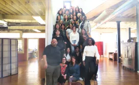 Large, diverse group of people standing and sitting on a wooden staircase in a bright office, smiling toward the camera.