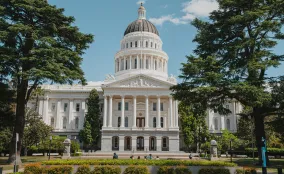 California State Capitol building in Sacramento framed by tall trees, with landscaped gardens and a bright blue sky.