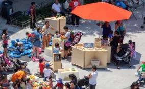Families and children gather around play tables and activity stations at an outdoor community plaza event.