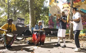 Community musicians play drums, congas, trumpet, and percussion in a sunny park beside a colorful mural.