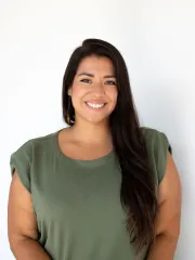 Smiling woman with long dark hair in a green top, posed against a bright white wall for a professional portrait.
