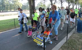 A group of community members, including adults and children, stand together outdoors holding handmade signs during a public gathering. The group appears to be participating in a community action or advocacy event in a park-like setting.