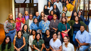 A large, diverse group of people poses together indoors, smiling warmly at the camera. They are gathered at the base of a staircase and across a brightly lit hallway, creating a vibrant and welcoming atmosphere. The group includes people of different ages, races, and genders, reflecting a strong sense of community and inclusion.