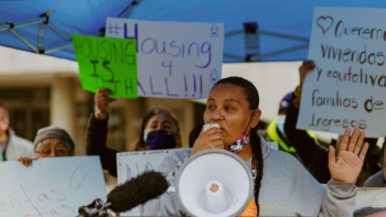 A woman speaks passionately into a megaphone at a housing justice rally, standing under a blue canopy. Behind her, people hold handmade signs with messages like “Housing 4 All!!!” and “Queremos viviendas equitativas.” The mood is one of empowerment and advocacy, as community members rally for affordable, equitable housing.