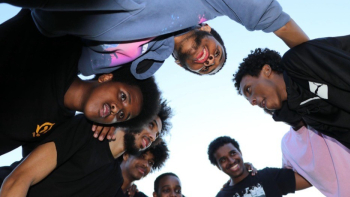 A group of young Black men huddle together in a close, supportive circle, looking down toward the camera with smiles and focused expressions. The sky is clear and blue behind them, creating a sense of unity, connection, and shared purpose.