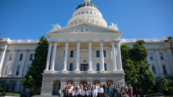 Large group of advocates, some in white coats, gathered on the steps of the California State Capitol beneath its dome.