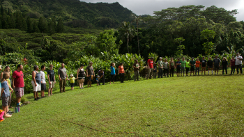 A large group of people stands together on a grassy field with trees and hills in the background.