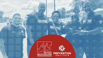 Group of community members at outdoor event wearing Aim4Peace shirts, with Prevention Institute and Big Cities Health Coalition branding