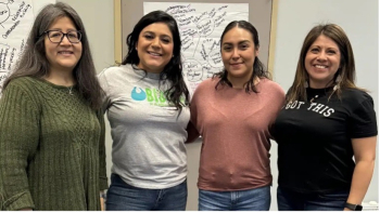Four women stand together smiling at the camera during a group gathering, with handwritten posters on the wall behind them.