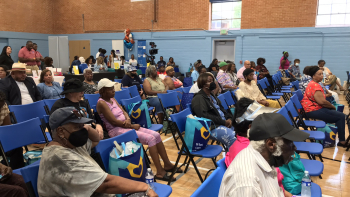 A community event taking place in a gymnasium with rows of seated attendees. Most individuals are older adults, and several are wearing masks. The attendees sit on blue chairs, some holding bags with health-related logos. A table in the back features informational materials, and staff are seen standing in the background. The setting is bright, with natural light coming through large windows.