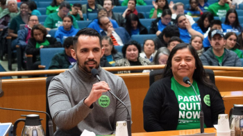 Man speaks into microphone while seated beside smiling woman in green shirt; audience fills room behind them at public event.