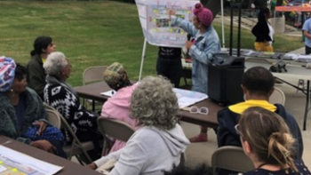  The image depicts a community gathering or public meeting taking place outdoors. A group of people, varying in age and likely representing a cross-section of the local community, are seated at tables and focused on a presentation. A person stands in the background under a tent, gesturing towards a display board that appears to contain maps or diagrams, possibly related to community planning or a local initiative. The scene is indicative of civic engagement.