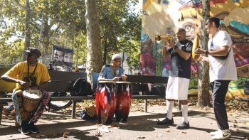 Community musicians play drums, congas, trumpet, and percussion in a sunny park beside a colorful mural.