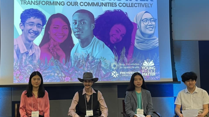 The image shows four young adults seated in front of a large projection screen displaying the words "YOUNG MINDS MATTER - TRANSFORMING OUR COMMUNITIES COLLECTIVELY." The backdrop features colorful portraits of diverse young individuals, aligning with the event's theme. The first person on the left is a woman wearing a pink blouse, the second person is a man with a mustache and a hat, the third is a woman in a gray blazer, and the fourth is a man in a light-colored shirt. 