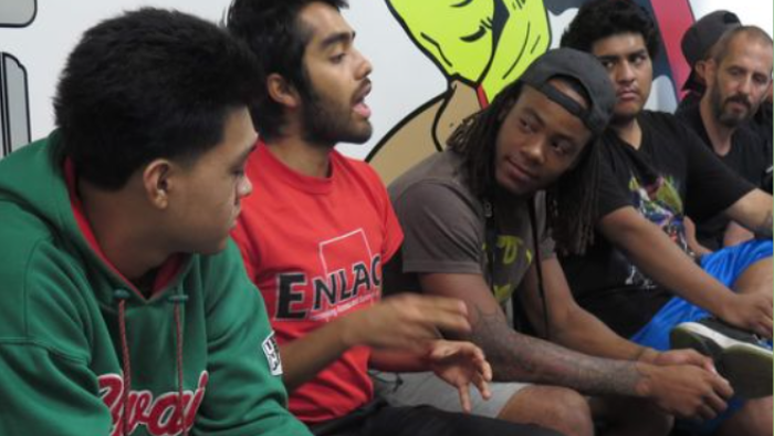 The image depicts a group of young men sitting and engaging in a discussion. The man in the foreground is wearing a green hoodie with the word "Coca-Cola" in white lettering. Beside him, a man in a red shirt with the text "ENLACE" is animatedly speaking and gesturing with his hand, indicating an active conversation. A man with dreadlocks, wearing a grey sleeveless shirt, is listening intently, and another in a blue shirt appears to be observing the discussion. 