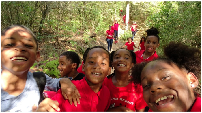 Smiling children in red shirts pose on a sunny forest trail, with others walking behind them among green trees.