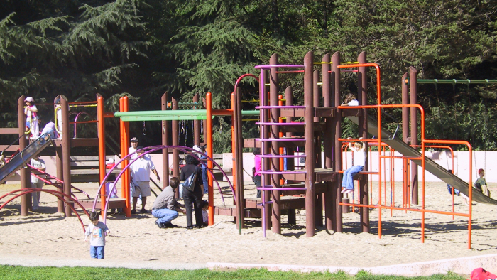 Children and caregivers play on a colorful playground structure with slides and climbing bars, surrounded by trees and sand.