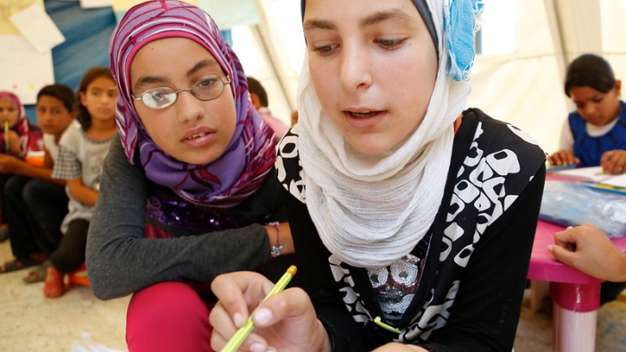The image shows two young girls engaged in an educational activity. The girl in the foreground is wearing a vibrant purple and pink hijab and glasses, and is attentively watching as the girl in the background, wearing a white and blue hijab, writes in a notebook. They both appear to be in a classroom setting, indicated by other children and educational materials in the background. The focus on the two girls suggests a learning environment that fosters interaction and guidance.