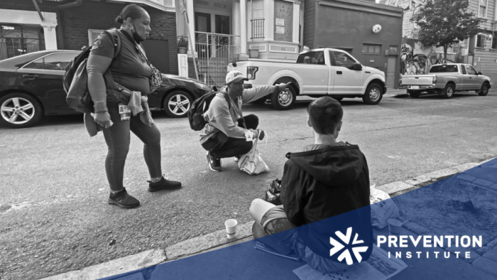 Outreach workers speak with a young person seated on a city sidewalk during a street-based community health outreach visit.