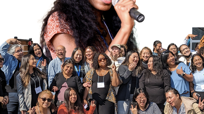 Speaker holding a microphone above a joyful, diverse group posing together outdoors, smiling, laughing, and celebrating community.